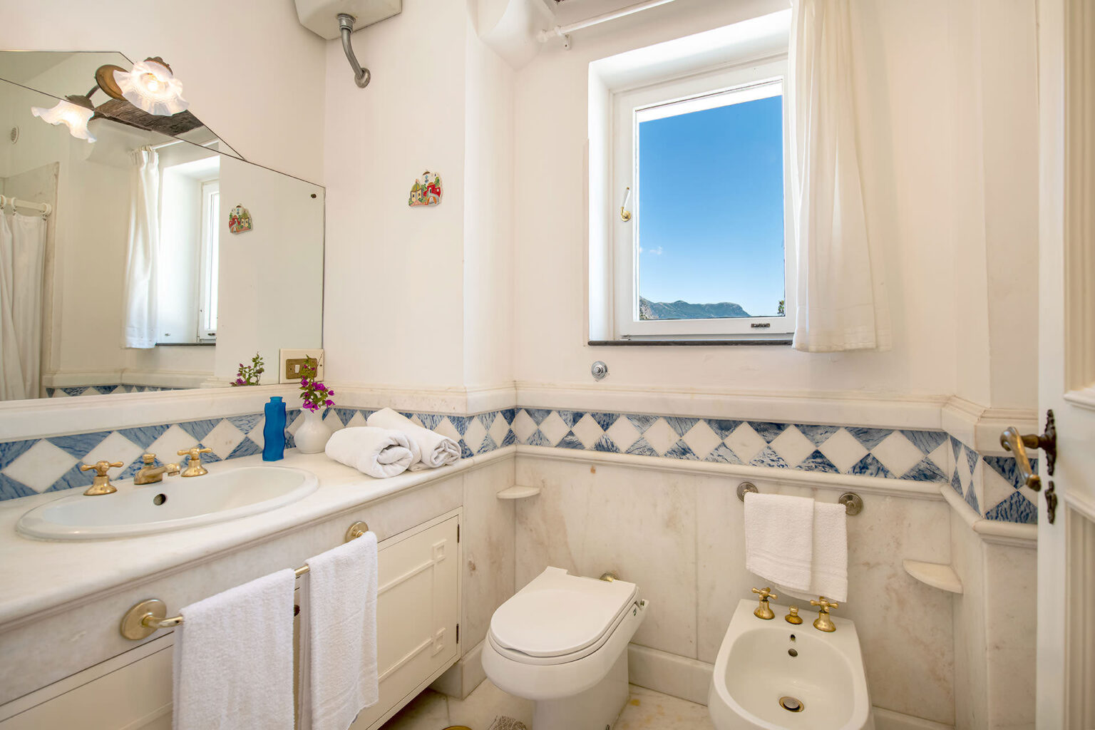 Bright and airy bathroom featuring a white vanity, blue and white tiled accents, a window with a scenic view, and neatly arranged towels. | Only on SopranoVillas