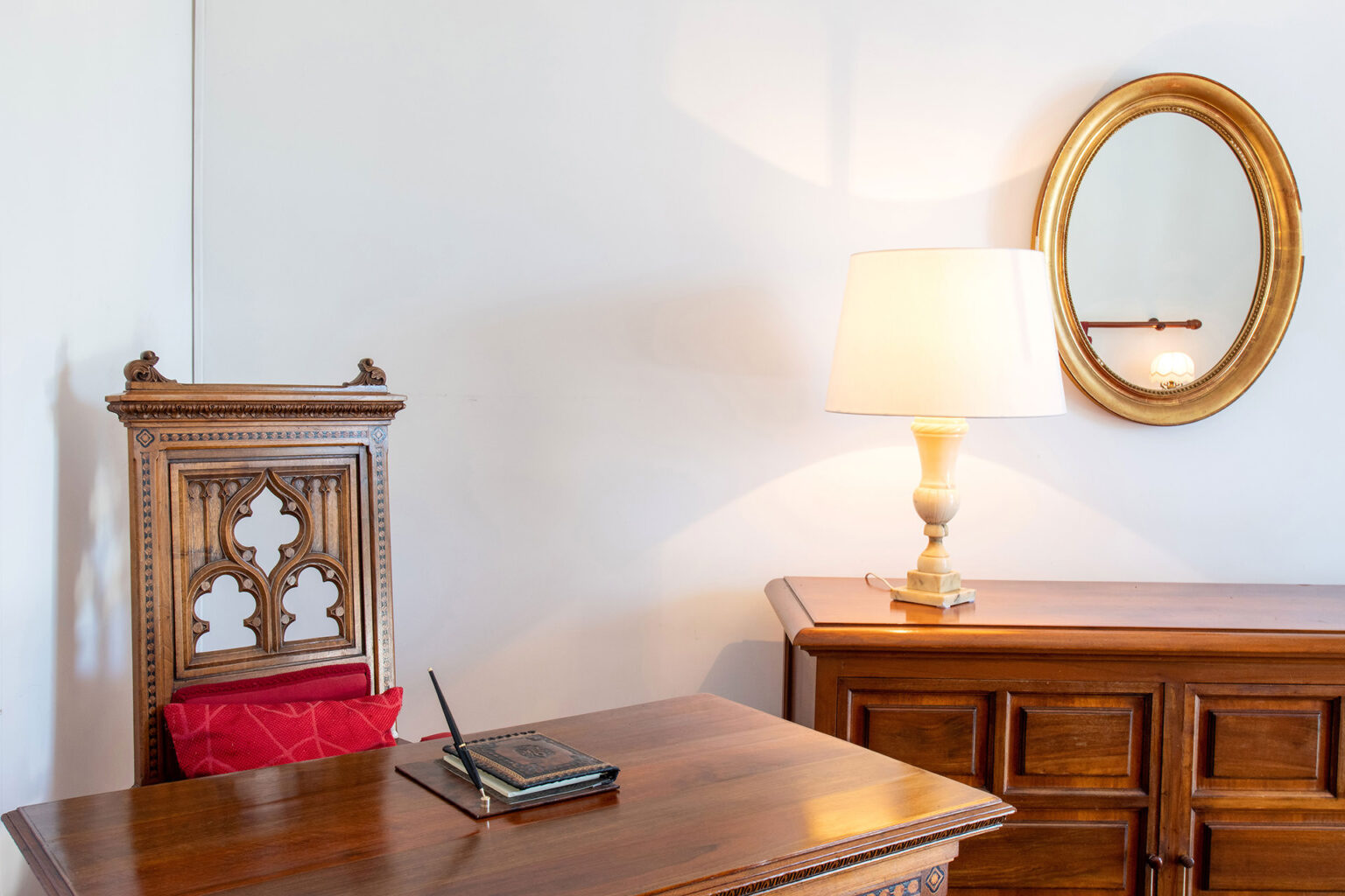 Antique wooden chair with intricate Gothic carvings, a desk with a notebook and pen, a stylish lamp, and a round gold-framed mirror in a well-lit room. | Only on SopranoVillas