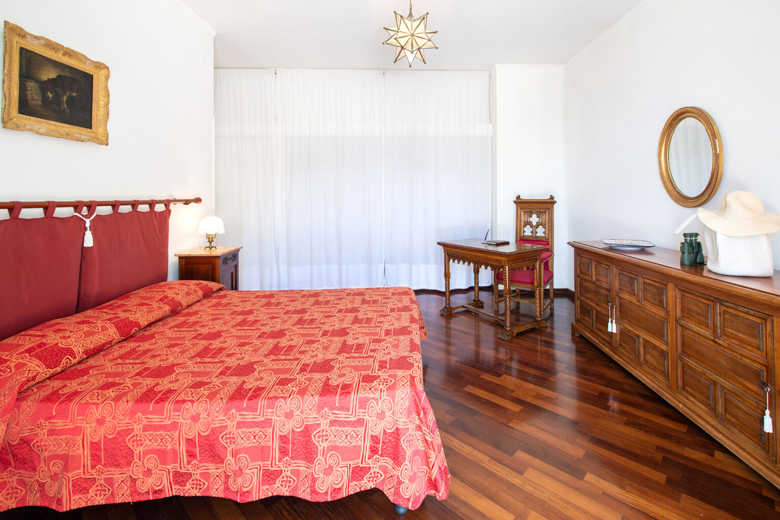 Cozy bedroom featuring a red patterned bedspread, wooden furniture, and a decorative painting. The room includes a bedside lamp, a vintage writing desk, and a stylish mirror, all set against a backdrop of bright white curtains and polished wooden flooring. | Only on SopranoVillas