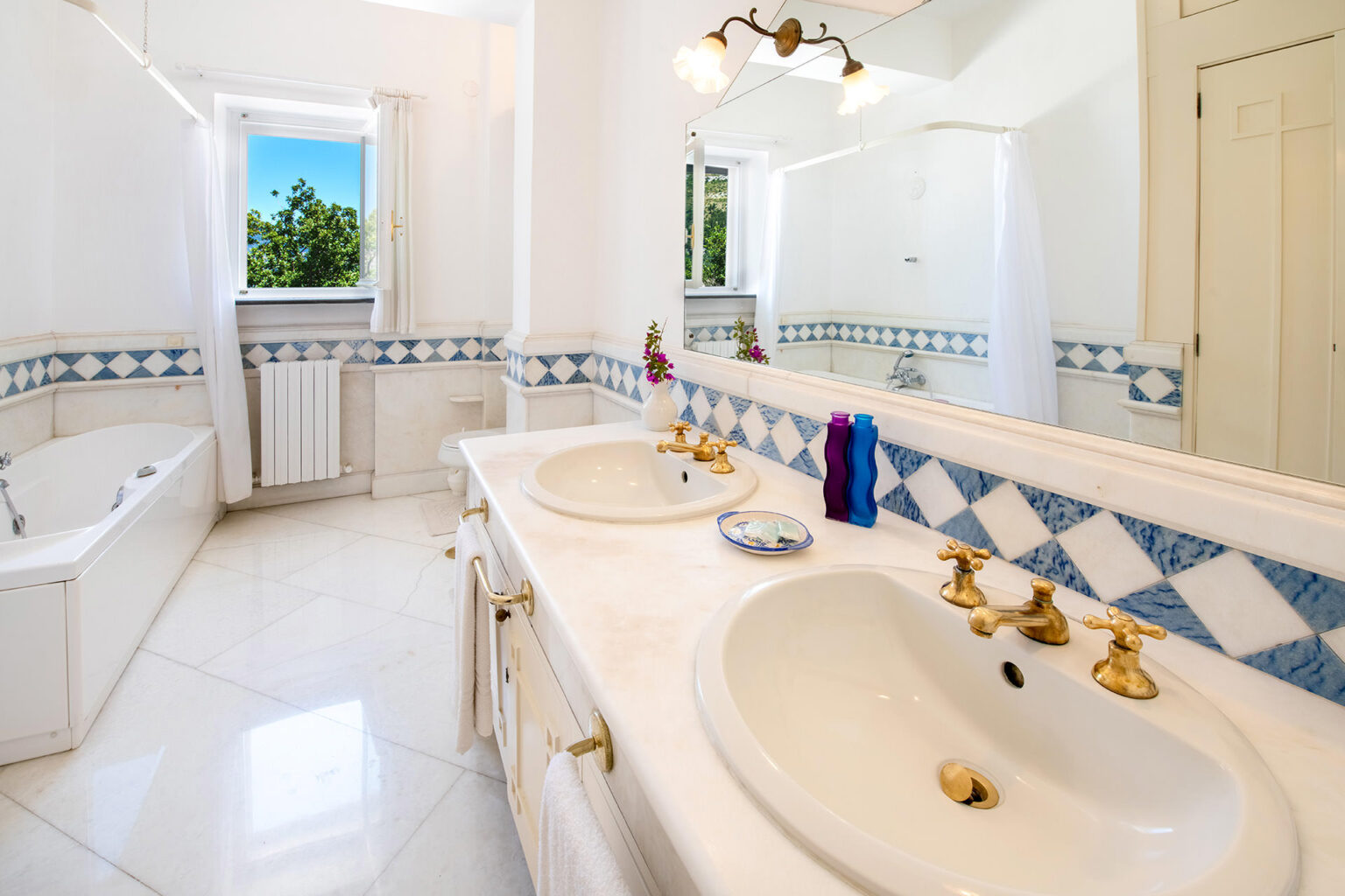 Bright and spacious bathroom featuring double sinks with gold fixtures, a large bathtub, and blue and white tiled accents. Natural light floods the room through a window, enhancing the clean, modern aesthetic. | Only on SopranoVillas