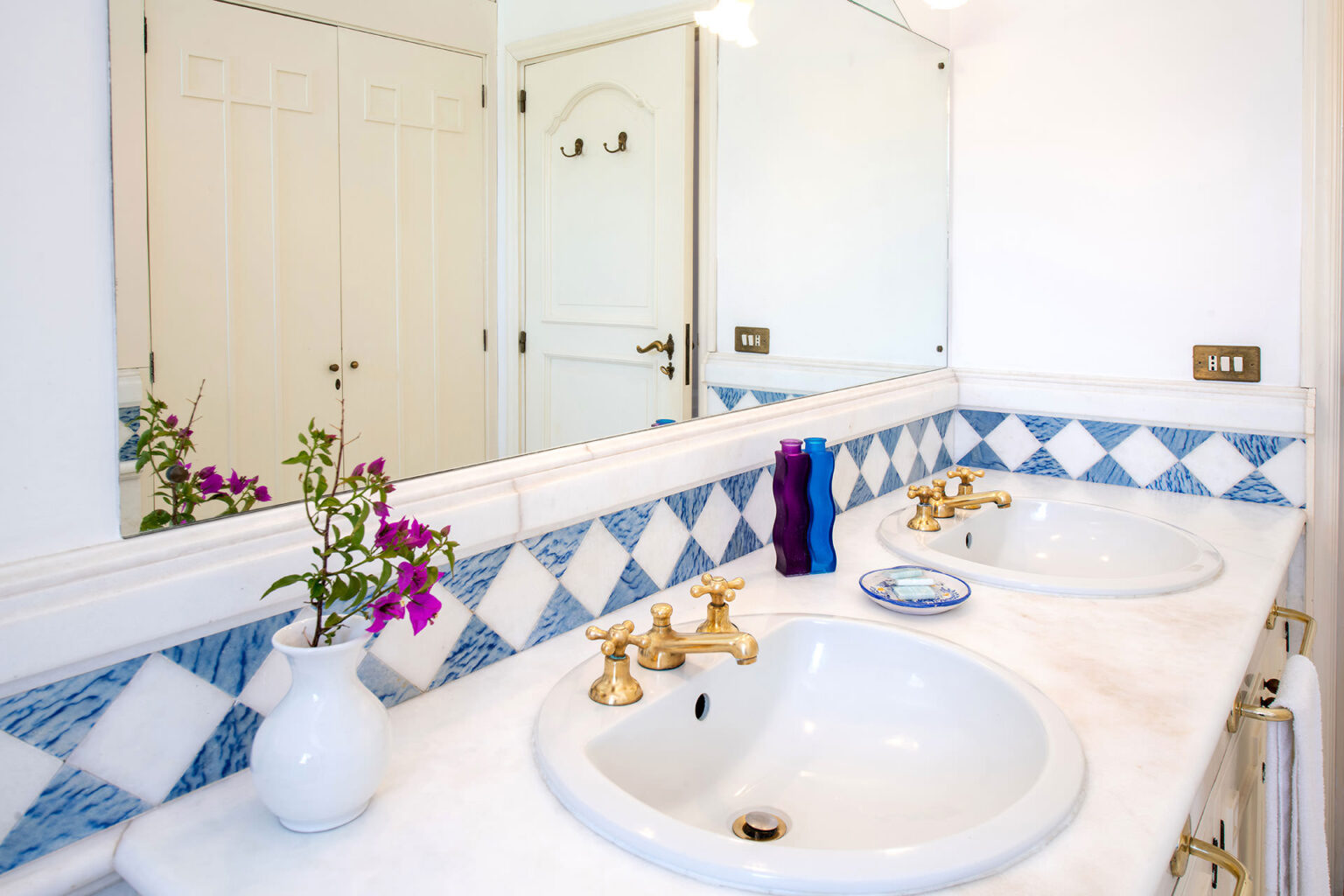 Bright and modern bathroom featuring dual sinks with elegant gold fixtures, a marble countertop, and a decorative blue and white tile backsplash. A small vase with purple flowers adds a touch of color, while a mirror reflects the space. | Only on SopranoVillas