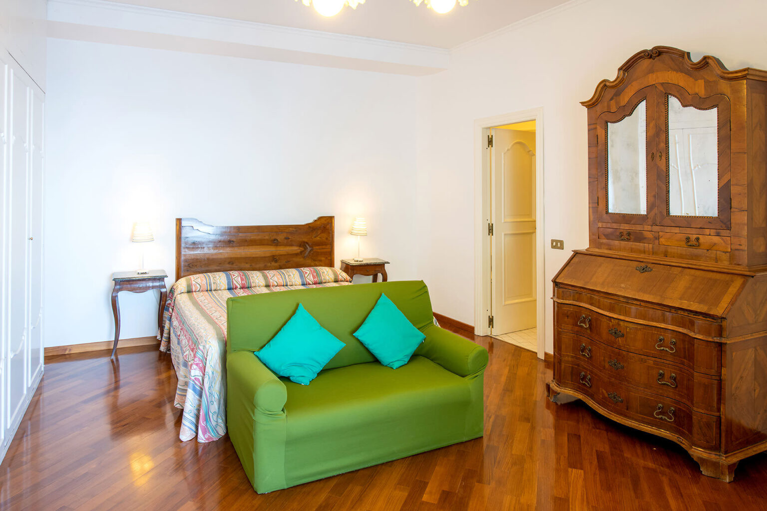 Cozy bedroom featuring a wooden bed with a colorful quilt, a green sofa with blue cushions, and an antique wooden dresser. The room has hardwood flooring and a bright, airy atmosphere. | Only on SopranoVillas