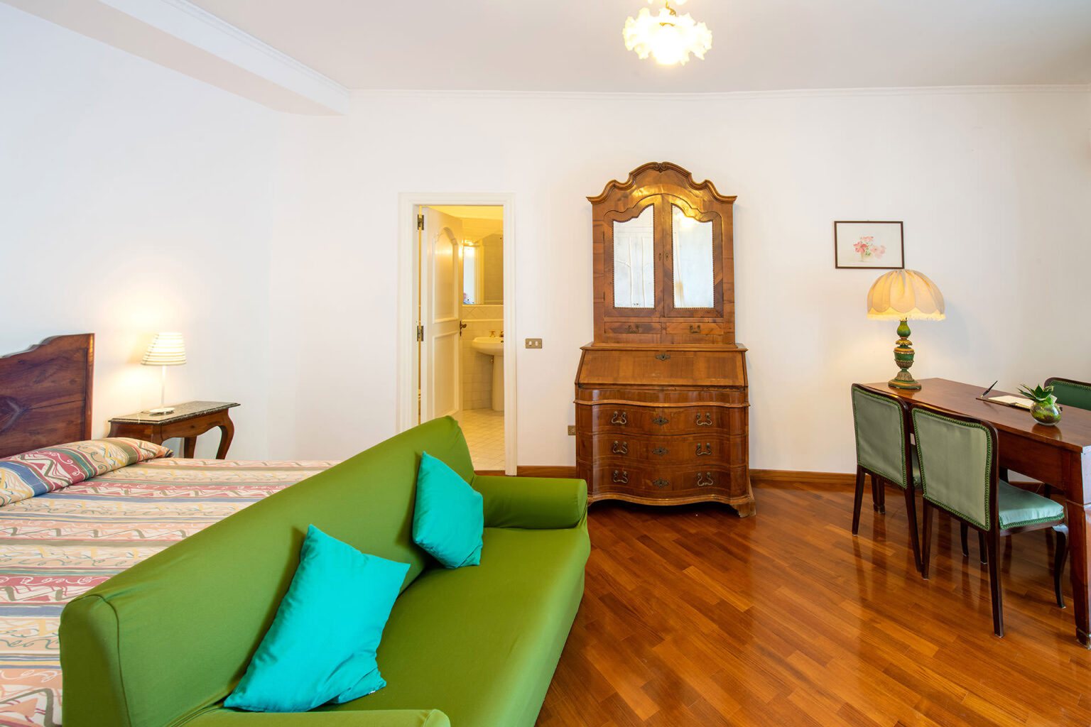 Cozy hotel room featuring a green sofa with decorative cushions, a wooden dresser, a dining table with chairs, and warm wooden flooring, creating an inviting atmosphere. | Only on SopranoVillas