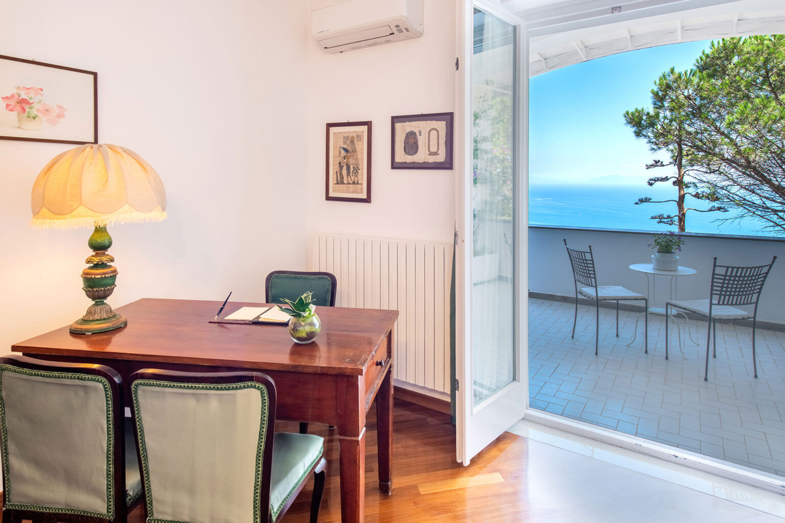 Cozy interior of a sunlit room featuring a wooden desk and green chairs, with a view of a balcony overlooking the sea and lush greenery. Ideal for relaxation and work. | Only on SopranoVillas