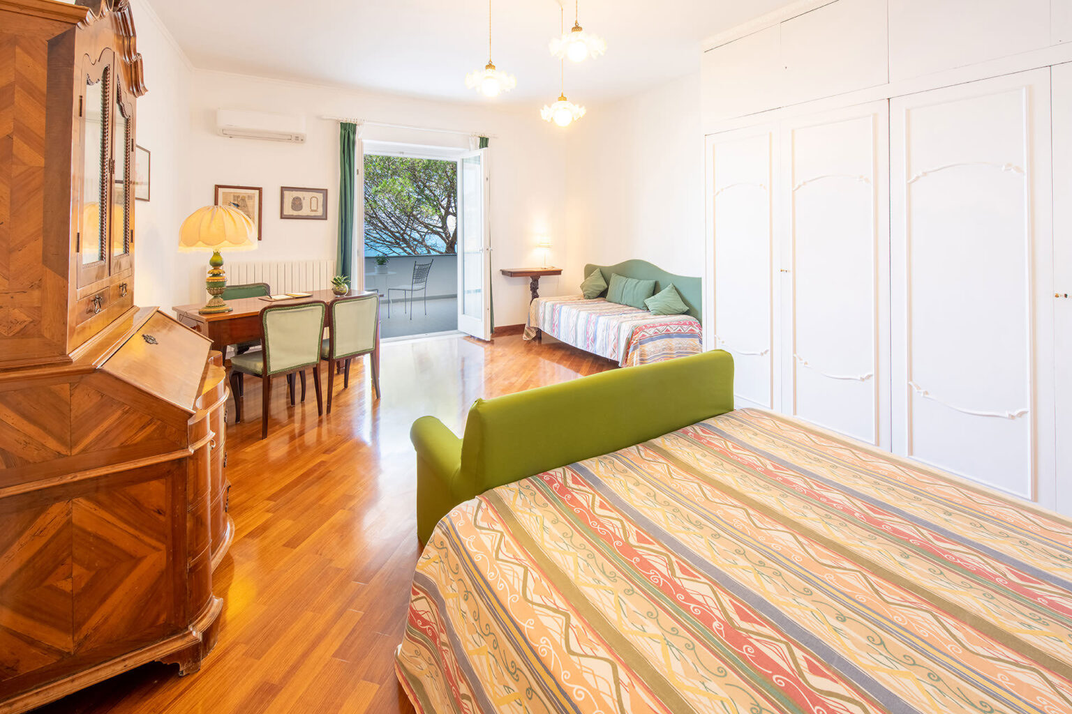 Bright and spacious bedroom featuring wooden flooring, a green bed, a wooden desk with chairs, and large windows overlooking a balcony. Decorated with warm colors and patterned bedding, the room combines comfort and elegance. | Only on SopranoVillas