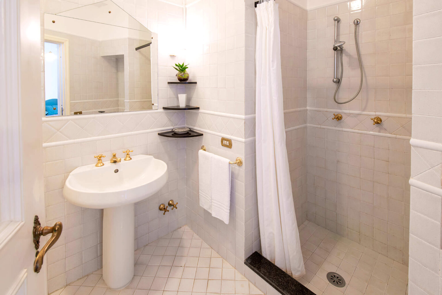 Modern bathroom featuring a walk-in shower, white tiled walls, a pedestal sink, and stylish fixtures. The space includes a small shelf with decorative plants and a towel rack for added convenience. | Only on SopranoVillas