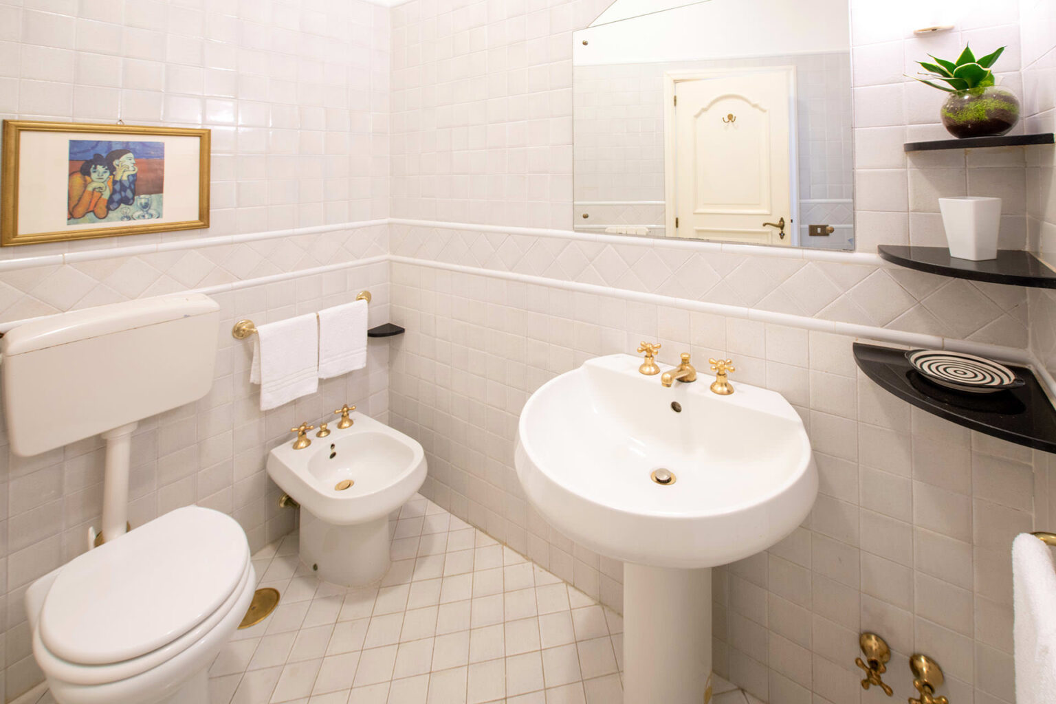 Modern bathroom featuring a white toilet, bidet, and sink with gold fixtures. The space is decorated with light tiles and a small plant on a black shelf, creating a clean and elegant aesthetic. | Only on SopranoVillas