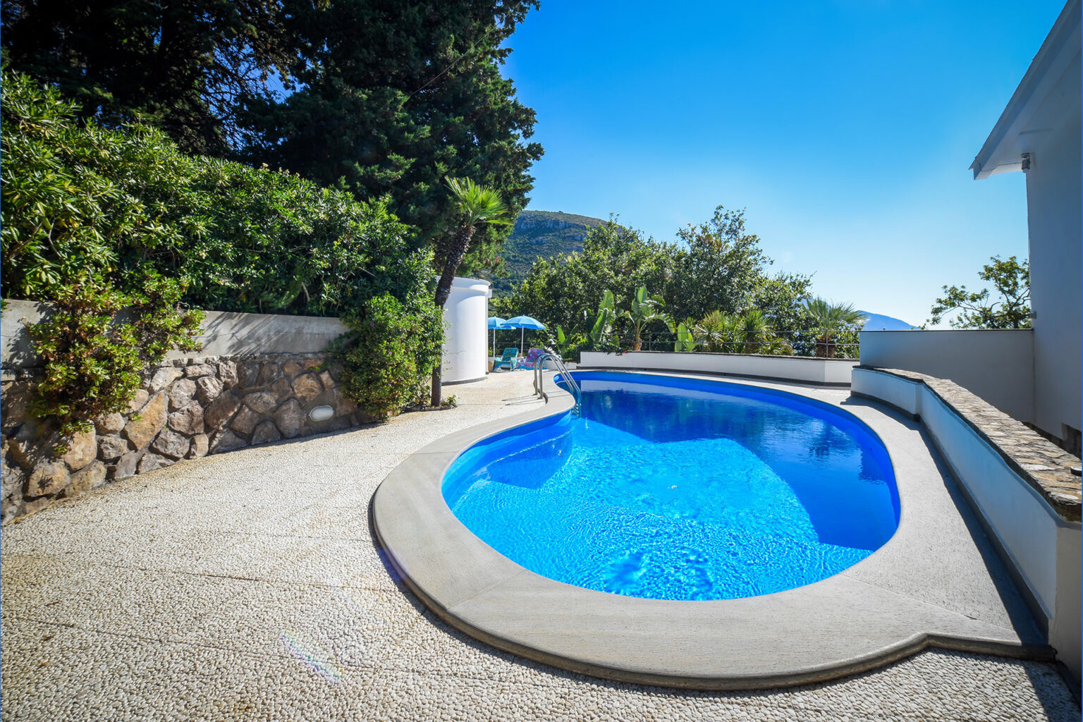 Luxury swimming pool surrounded by lush greenery and stone wall, featuring a clear blue water surface under a sunny sky, ideal for relaxation and outdoor leisure. | Only on SopranoVillas