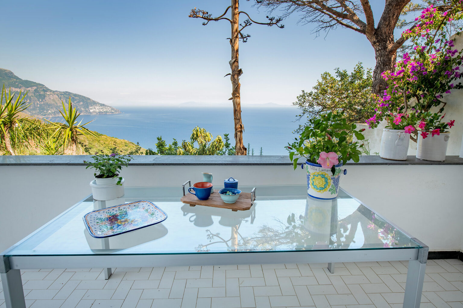 Glass table set on a terrace overlooking a serene ocean view, adorned with colorful potted plants and tableware, ideal for outdoor dining and relaxation. | Only on SopranoVillas