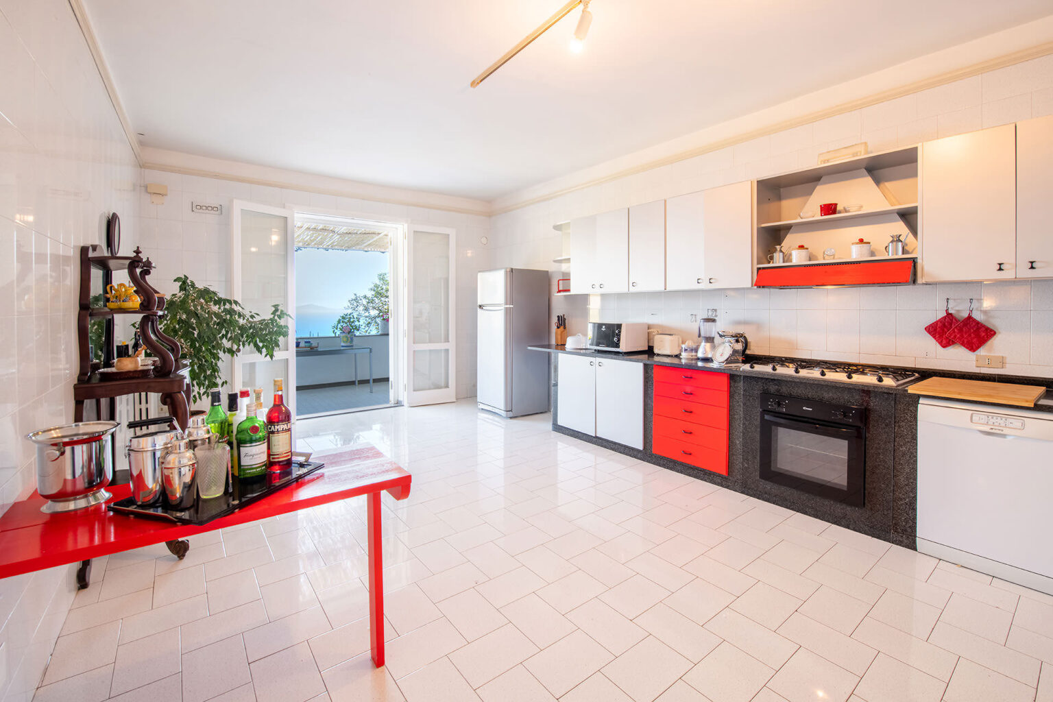 Bright and modern kitchen featuring white and red cabinetry, stainless steel appliances, and a spacious layout with a door leading to a scenic outdoor area. The kitchen includes a dining table with a bar setup and a view of nature outside. | Only on SopranoVillas