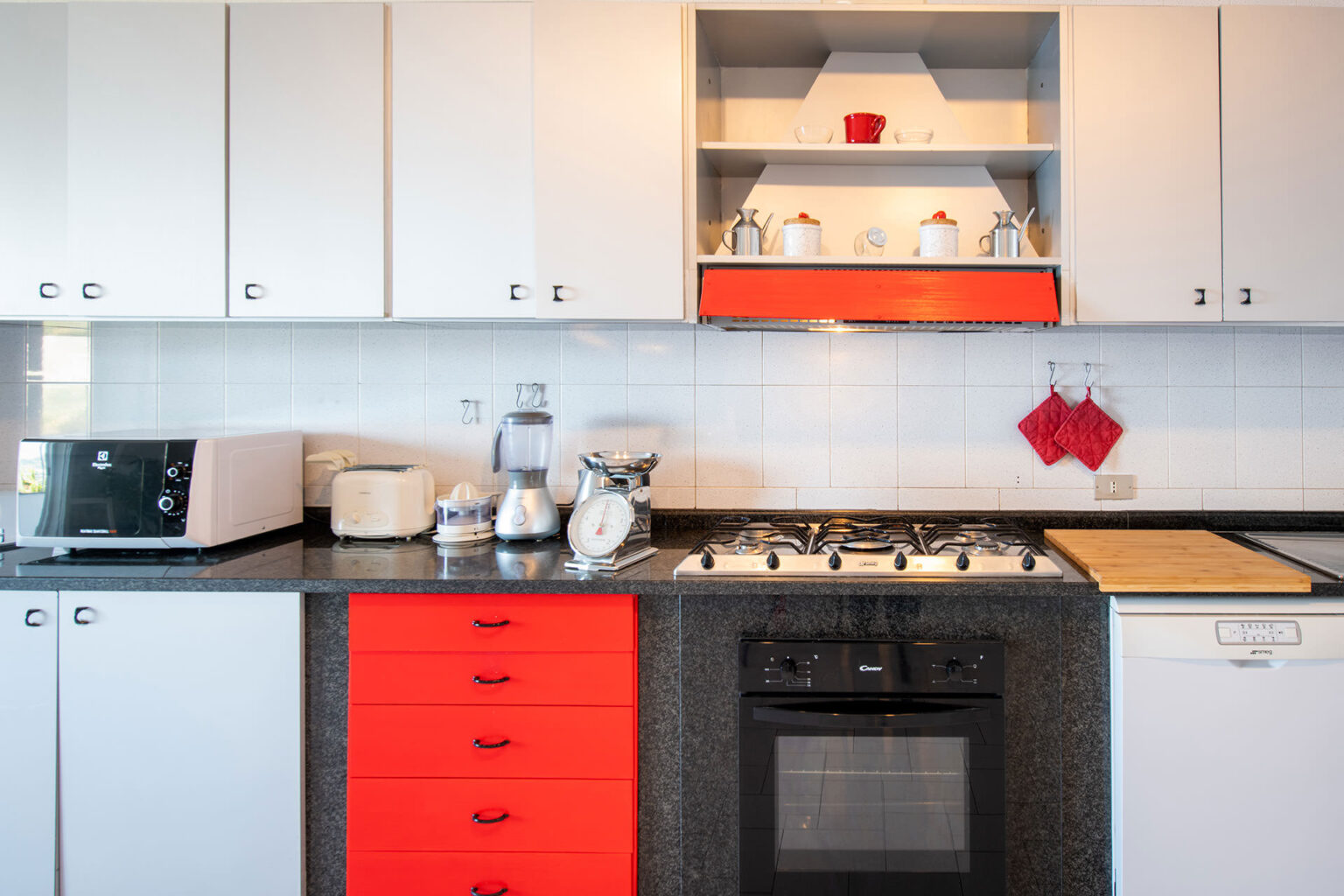Modern kitchen featuring white cabinets with red accents, a black countertop, and essential appliances including a microwave, toaster, blender, and gas stove. The design combines functionality with a vibrant color scheme, showcasing an organized layout with hanging pot holders and a wooden cutting board. | Only on SopranoVillas