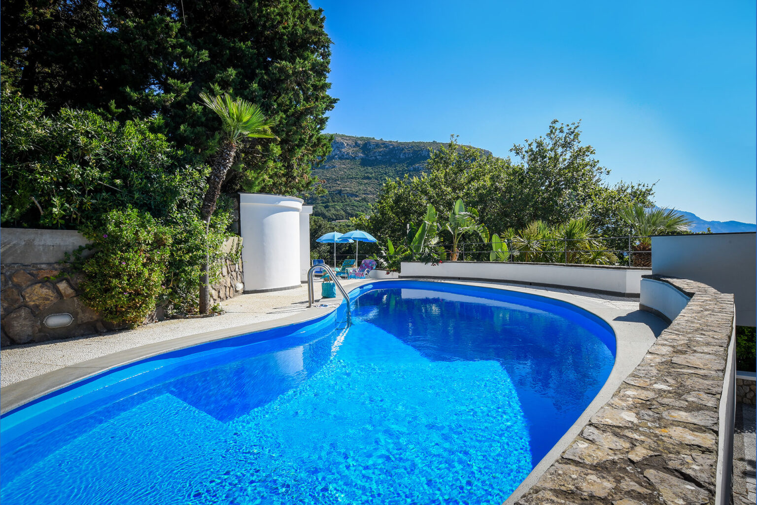 Scenic outdoor swimming pool surrounded by lush greenery, stone walls, and a clear blue sky, ideal for relaxation and leisure activities. | Only on SopranoVillas