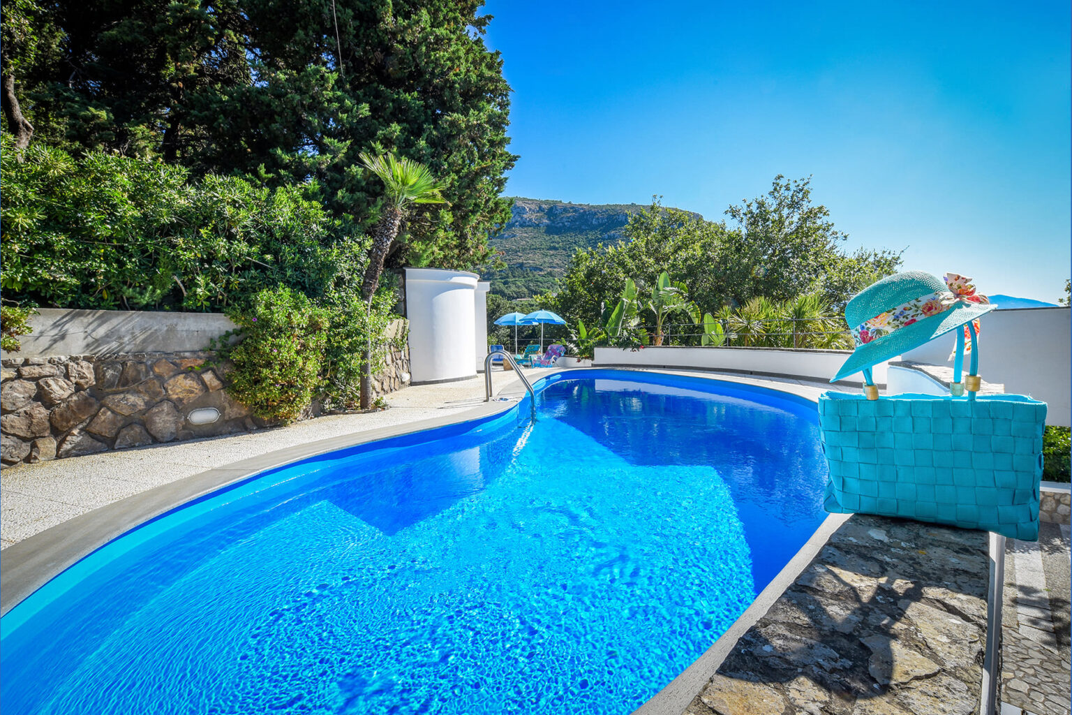 Vibrant outdoor swimming pool surrounded by lush greenery and stone walls, featuring a bright blue water surface, a stylish turquoise sun hat, and a matching tote bag, set against a clear blue sky and a scenic mountain backdrop. | Only on SopranoVillas