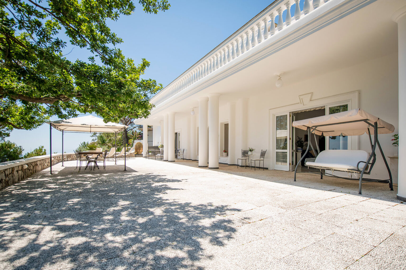 Spacious outdoor terrace of a modern villa featuring white columns, a shaded dining area with a table and chairs, and a relaxing swing, surrounded by lush greenery and blue skies. | Only on SopranoVillas