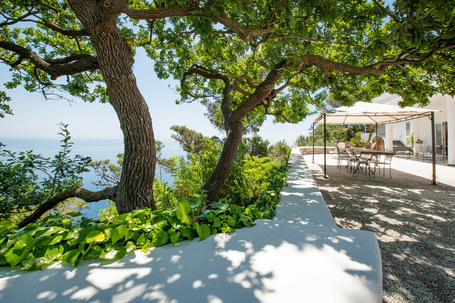 Scenic outdoor patio area featuring lush green trees and a view of the ocean, with a shaded dining space and modern furniture, perfect for relaxation and entertaining. | Only on SopranoVillas