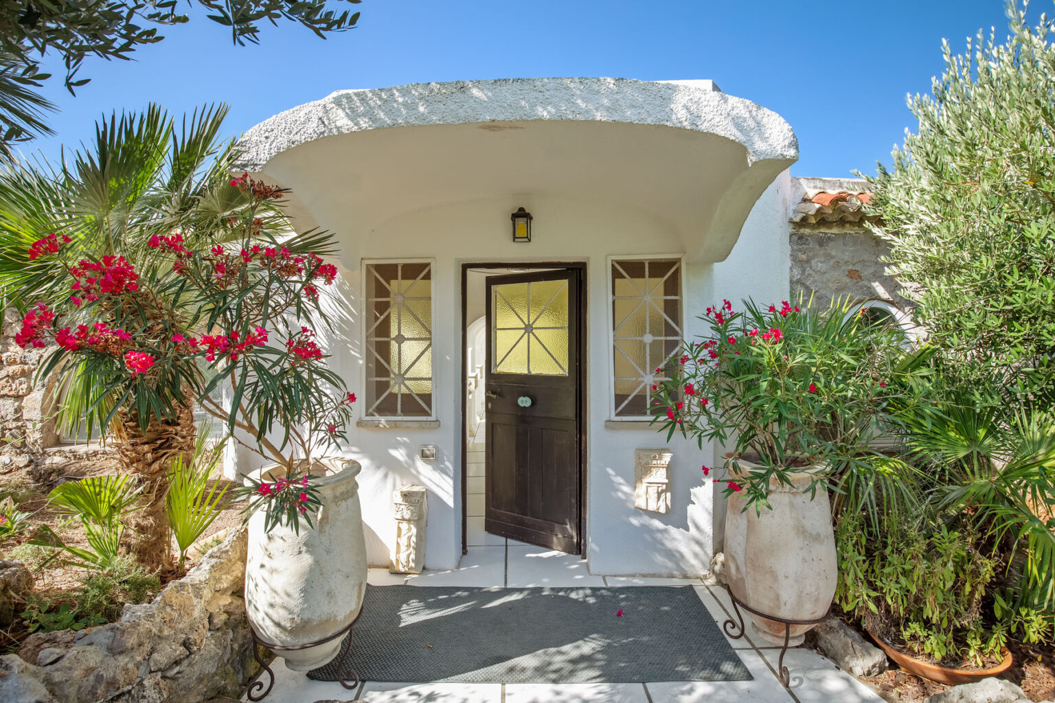 Charming entrance of a Mediterranean-style villa featuring a white stone facade, wooden door, and vibrant flowering plants in decorative pots, surrounded by lush greenery under a clear blue sky. | Only on SopranoVillas