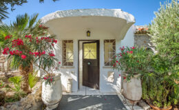 Charming entrance of a Mediterranean-style villa featuring a white stone facade, wooden door, and vibrant flowering plants in decorative pots, surrounded by lush greenery under a clear blue sky.
