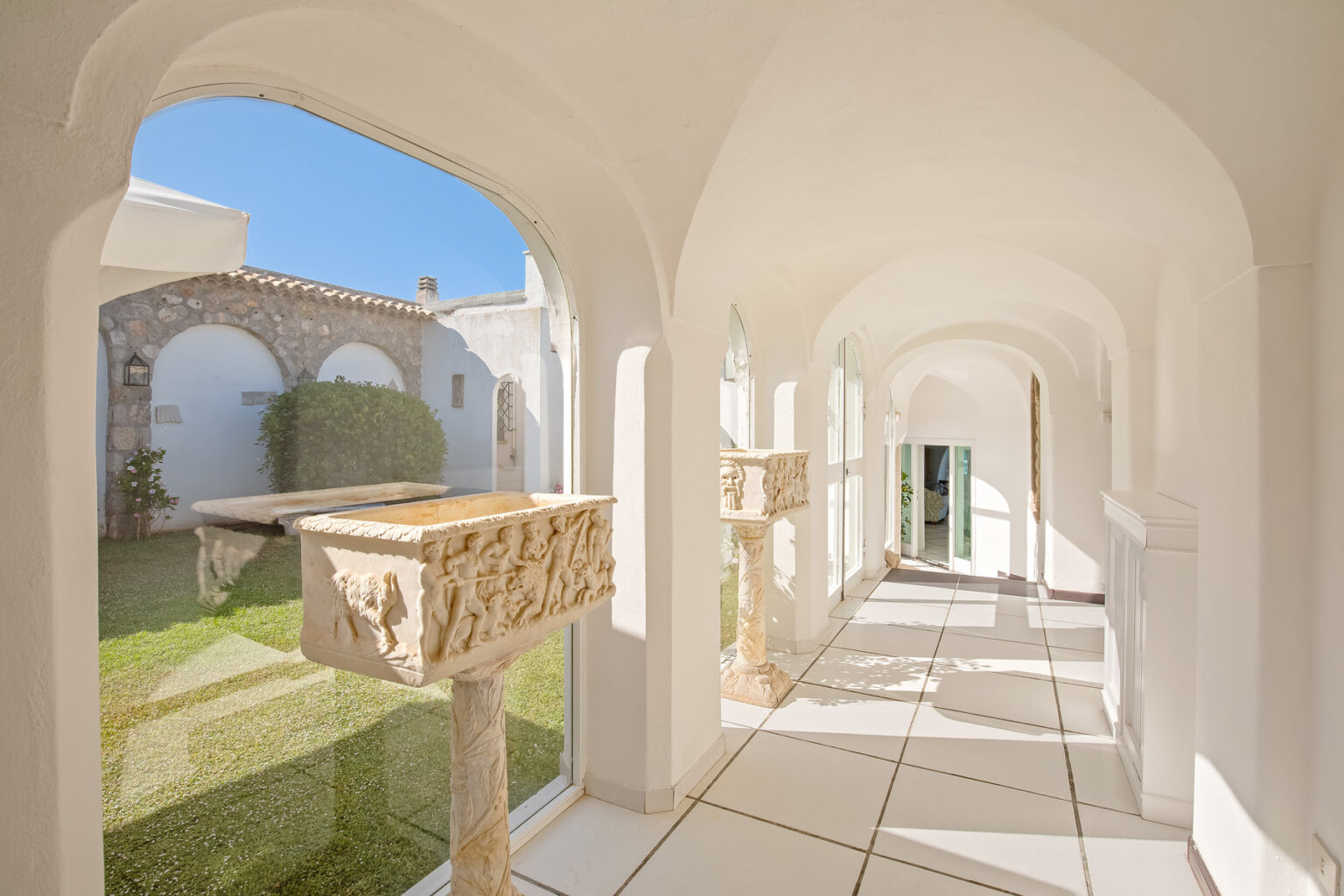 Bright and airy corridor with arched windows, featuring stone planters and views of a landscaped garden, showcasing modern architectural design and natural light. | Only on SopranoVillas