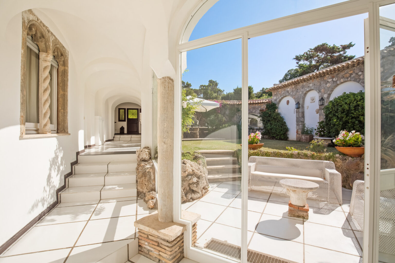 Bright and airy interior view of a modern hallway leading to a landscaped garden, featuring large glass doors, white tiled floors, and decorative stone elements. | Only on SopranoVillas