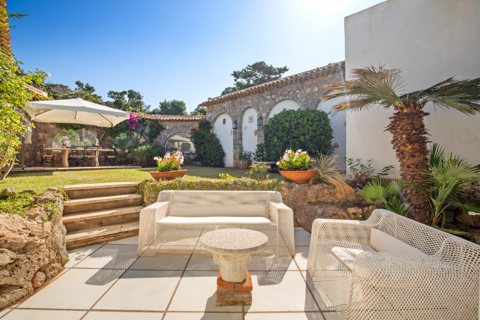 Cozy outdoor seating area with white wicker furniture, surrounded by lush greenery and vibrant flowers, featuring stone steps leading to a beautifully landscaped garden under a clear blue sky. | Only on SopranoVillas
