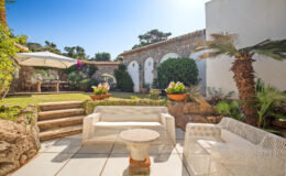 Cozy outdoor seating area with white wicker furniture, surrounded by lush greenery and vibrant flowers, featuring stone steps leading to a beautifully landscaped garden under a clear blue sky.