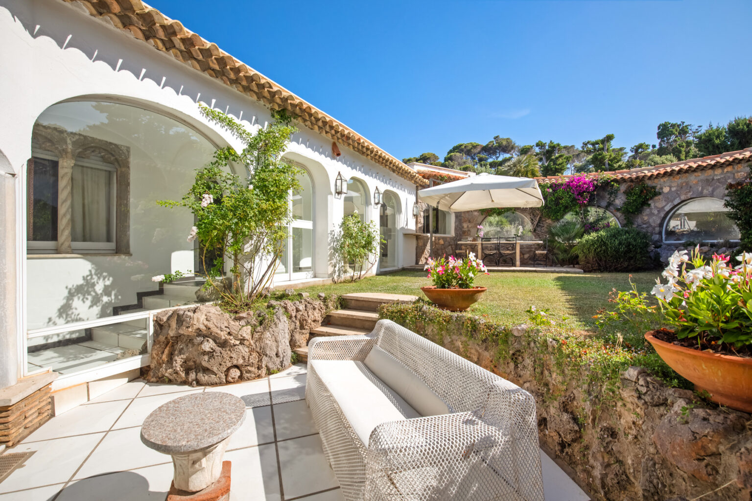 Charming outdoor patio featuring a modern white sofa, stone accents, and lush greenery, set against a backdrop of a beautifully designed house with arched windows and a sunny blue sky. | Only on SopranoVillas