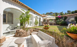 Charming outdoor patio featuring a modern white sofa, stone accents, and lush greenery, set against a backdrop of a beautifully designed house with arched windows and a sunny blue sky.