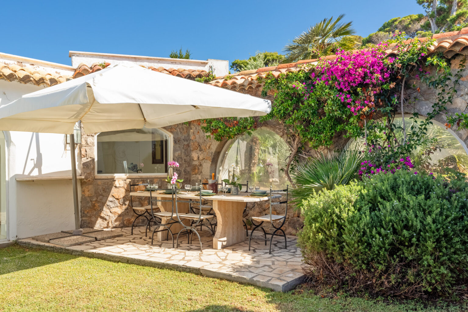 Outdoor dining area with a stone table and black chairs, shaded by a white umbrella, surrounded by lush greenery and vibrant flowers, perfect for al fresco meals in a sunny garden setting. | Only on SopranoVillas