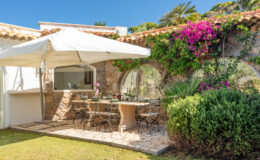 Outdoor dining area with a stone table and black chairs, shaded by a white umbrella, surrounded by lush greenery and vibrant flowers, perfect for al fresco meals in a sunny garden setting.