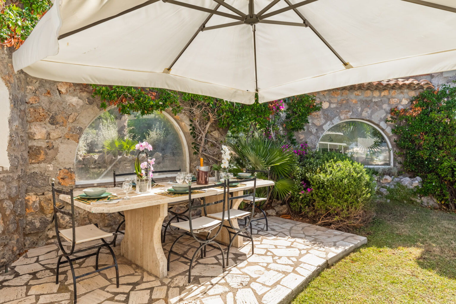 Outdoor dining area featuring a stone table set with plates, glasses, and a floral centerpiece, shaded by a large umbrella. Lush greenery and colorful flowers surround the space, creating a serene atmosphere. | Only on SopranoVillas