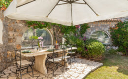 Outdoor dining area featuring a stone table set with plates, glasses, and a floral centerpiece, shaded by a large umbrella. Lush greenery and colorful flowers surround the space, creating a serene atmosphere.