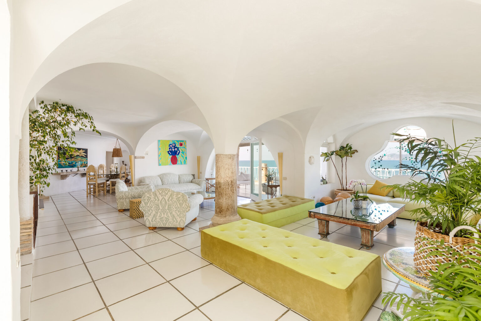Spacious and bright living room featuring arched ceilings, modern furniture, and large windows with ocean views, complemented by vibrant artwork and indoor plants. | Only on SopranoVillas