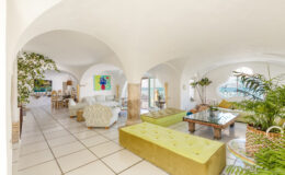 Spacious and bright living room featuring arched ceilings, modern furniture, and large windows with ocean views, complemented by vibrant artwork and indoor plants.