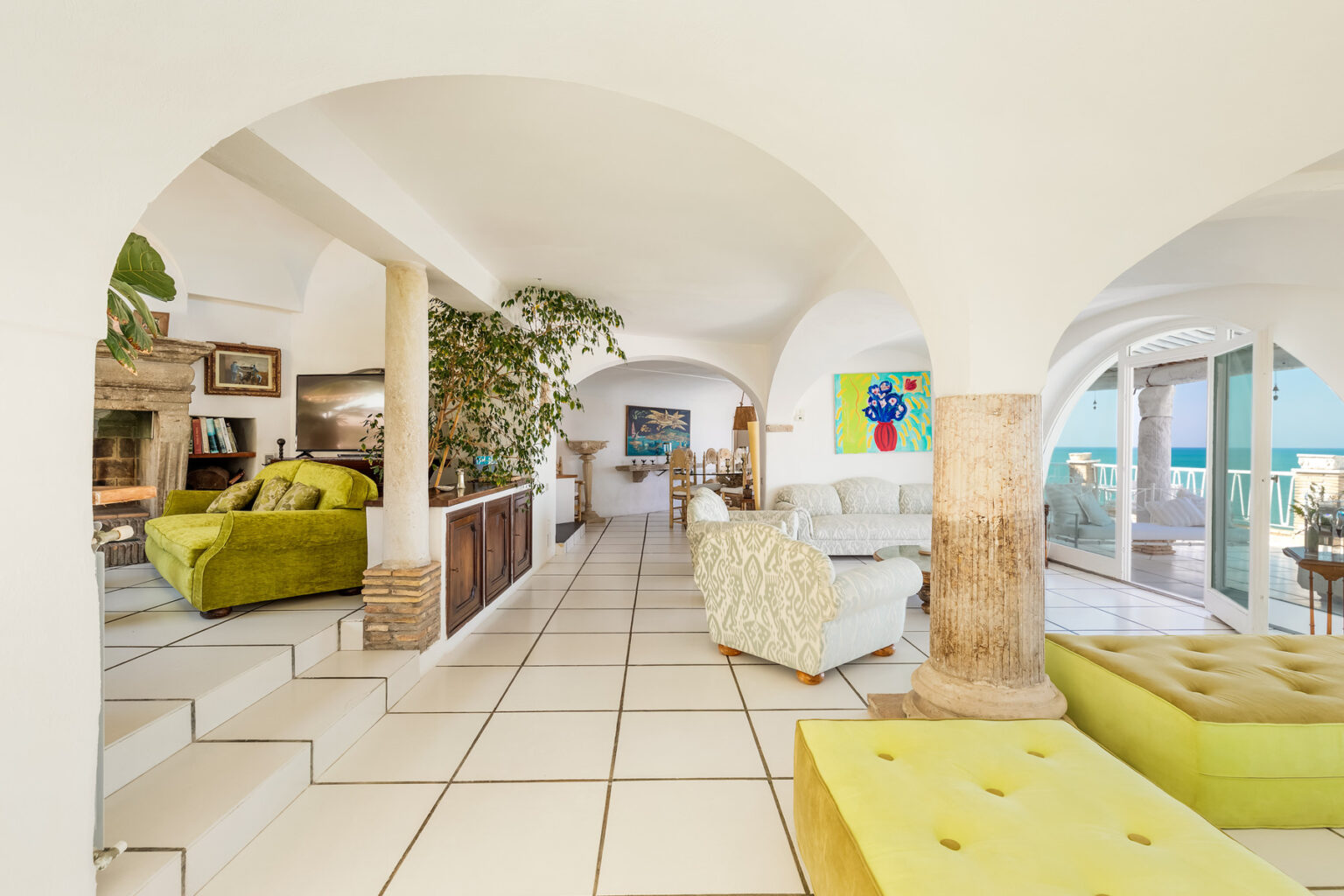 Bright and airy interior of a modern living room featuring arched doorways, vibrant green furniture, and large windows that offer ocean views. The space is elegantly decorated with plants and artwork, showcasing a blend of comfort and style. | Only on SopranoVillas