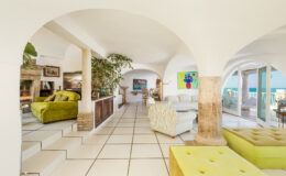 Bright and airy interior of a modern living room featuring arched doorways, vibrant green furniture, and large windows that offer ocean views. The space is elegantly decorated with plants and artwork, showcasing a blend of comfort and style.