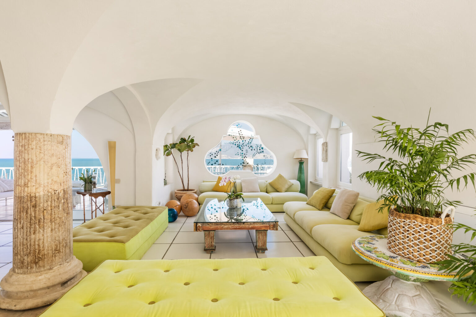 Bright and airy lounge area featuring modern green seating, decorative plants, and a stunning ocean view through an arched window, perfect for relaxation and social gatherings. | Only on SopranoVillas