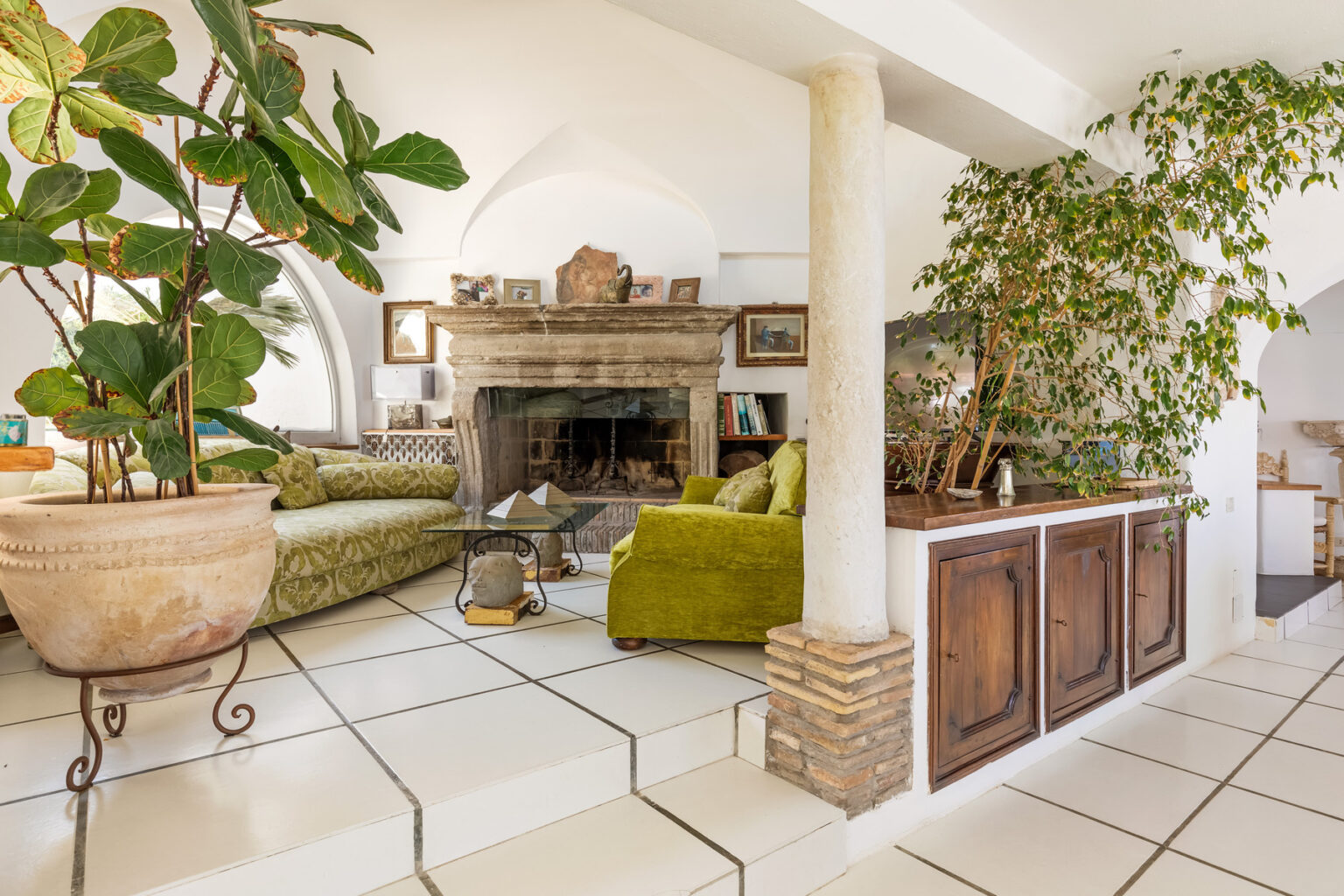 Cozy living room featuring a green sofa, a stone fireplace, and lush indoor plants, complemented by a tiled floor and wooden cabinetry. Ideal for a warm, inviting atmosphere. | Only on SopranoVillas
