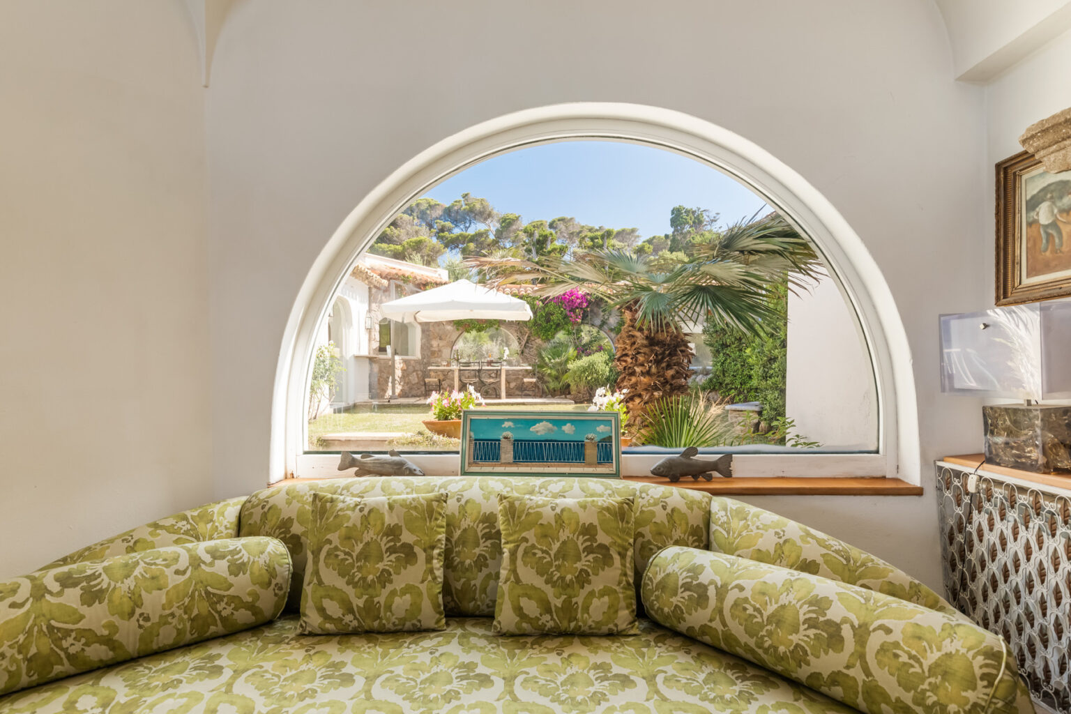 Cozy interior view featuring a curved window overlooking a vibrant garden with a patio, colorful furniture, and lush greenery, highlighting a serene outdoor space. | Only on SopranoVillas