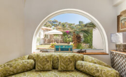 Cozy interior view featuring a curved window overlooking a vibrant garden with a patio, colorful furniture, and lush greenery, highlighting a serene outdoor space.