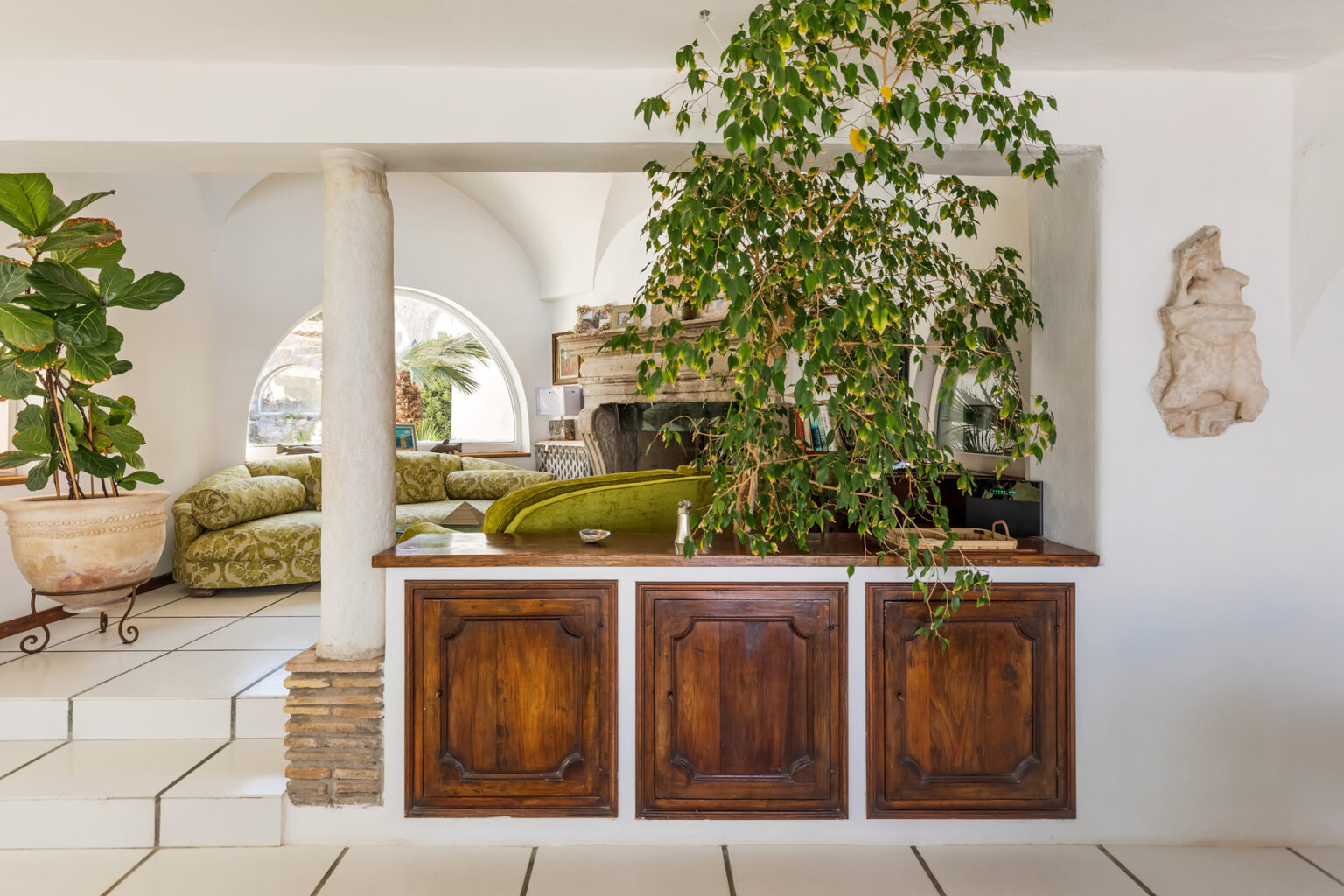 Interior view of a stylish living space featuring a wooden bar with decorative paneling, surrounded by lush greenery, and an inviting seating area with green upholstery. The space is bright and airy, showcasing a blend of modern and rustic design elements. | Only on SopranoVillas