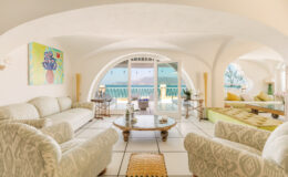 Spacious and elegantly designed living room featuring a light color palette, plush sofas, and natural light streaming through large arched windows. The room is adorned with contemporary art and tasteful décor, creating a warm and inviting atmosphere.