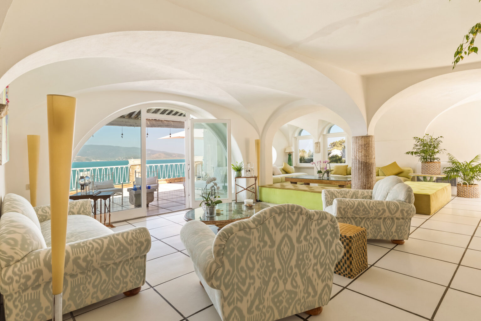 Bright and airy living room with elegant furniture, large windows offering a scenic view, and lush greenery, creating a serene coastal atmosphere. | Only on SopranoVillas