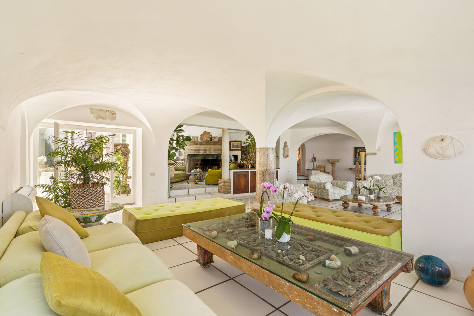 Spacious living room with arched doorways, featuring a cozy yellow sofa, a wooden coffee table adorned with decorative stones, and lush indoor plants, creating a bright and inviting atmosphere. | Only on SopranoVillas