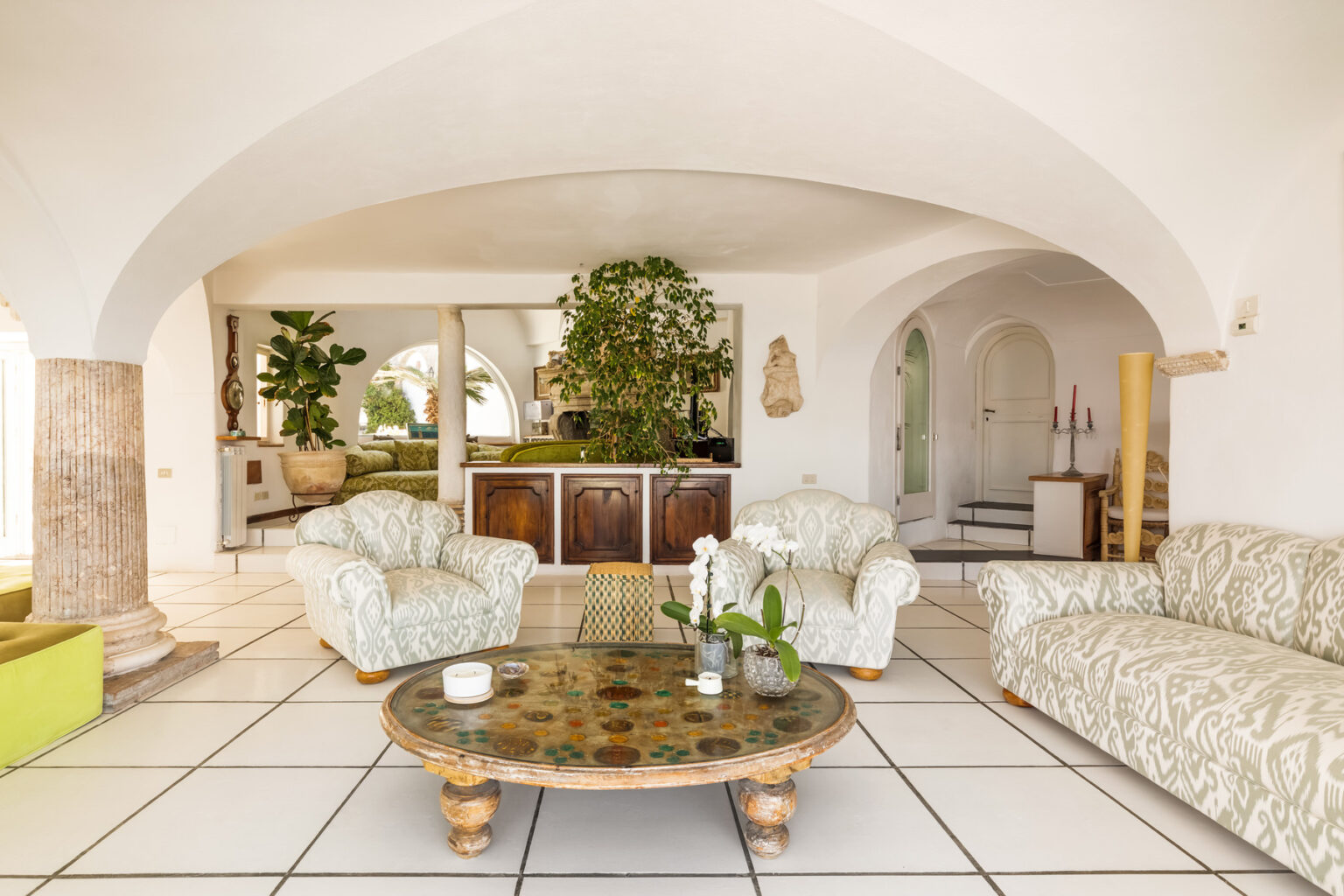 Spacious and bright living room featuring arched ceilings, comfortable patterned sofas, a decorative coffee table, and lush indoor plants, creating a serene and inviting atmosphere. | Only on SopranoVillas