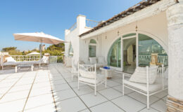 Spacious terrace of a modern villa featuring white outdoor furniture, a large umbrella for shade, and panoramic views under a clear blue sky. The design showcases elegant architectural details and a serene atmosphere, perfect for relaxation.
