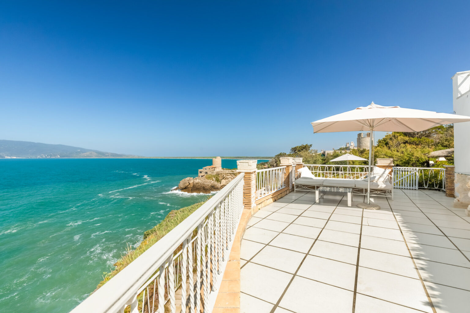 Scenic ocean view from a coastal balcony with a sun lounger and umbrella, showcasing clear blue skies and gentle waves. | Only on SopranoVillas