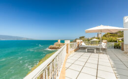 Scenic ocean view from a coastal balcony with a sun lounger and umbrella, showcasing clear blue skies and gentle waves.