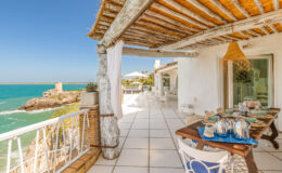 Cozy seaside terrace with a dining table set for guests, featuring ocean views and a rustic design. The space includes white furniture, a thatched roof, and a glimpse of a historical building on the rocky shore. Ideal for relaxation and outdoor dining.