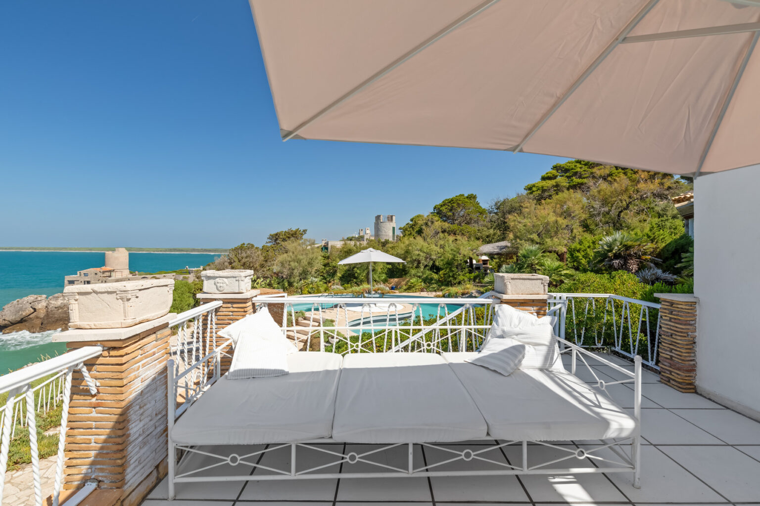Cozy outdoor lounge area with white cushions overlooking a serene ocean view, surrounded by lush greenery and a swimming pool, under a sunshade on a bright sunny day. | Only on SopranoVillas