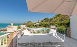 Cozy outdoor lounge area with white cushions overlooking a serene ocean view, surrounded by lush greenery and a swimming pool, under a sunshade on a bright sunny day.